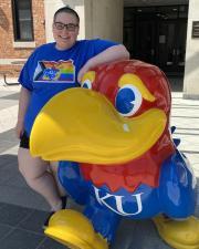 Cori Deming leaning against a Jayhawk statue while wearing a blue progress flag Jayhawk shirt.